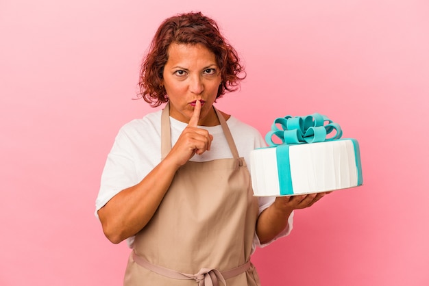 Pastelería de mediana edad mujer latina sosteniendo un pastel aislado sobre fondo rosa guardando un secreto o pidiendo silencio.