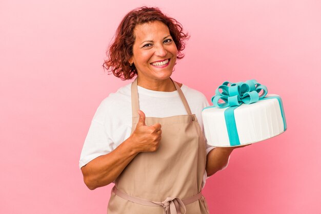 Pastelería de mediana edad mujer latina sosteniendo un pastel aislado sobre fondo rosa sonriendo y levantando el pulgar hacia arriba