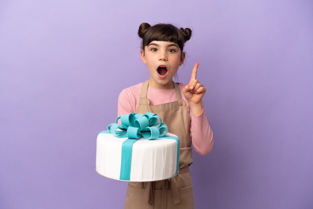 Pastelería niña sosteniendo un gran pastel aislado en la pared púrpura con la intención de darse cuenta de la solución mientras levanta un dedo