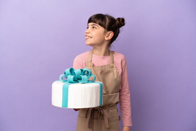 Pastelería niña sosteniendo un gran pastel aislado en la pared púrpura mirando hacia el lado y sonriendo