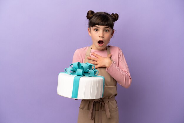 Pastelería niña sosteniendo un gran pastel aislado en la pared púrpura sorprendida y conmocionada mientras mira a la derecha