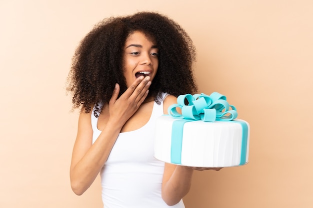 Pastelero afro chef sosteniendo un gran pastel aislado en beige con sorpresa y expresión facial conmocionada