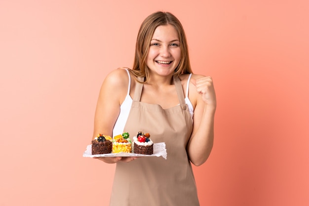 Pastelero chef ucraniano sosteniendo un muffins aislado en rosa espacio celebrando una victoria