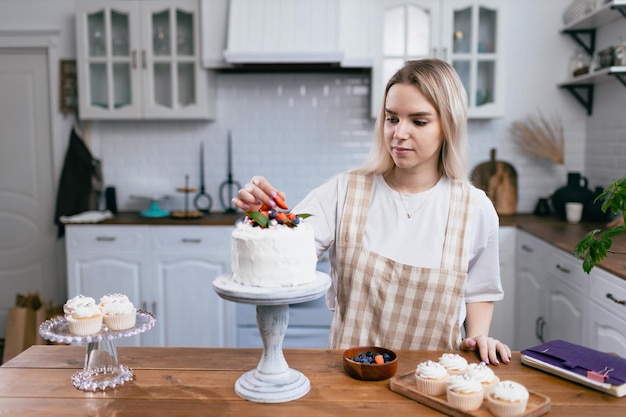 Pastelero confitero joven en delantal beige mujer decorar pastel en la mesa de la cocina Pasteles cupcakes y postre dulce