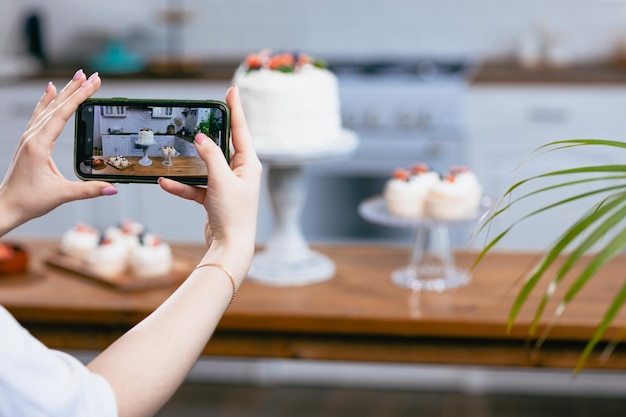 Pastelero confitero joven mujer caucásica tomar una foto en el teléfono celular pastel en la mesa de la cocina Pasteles cupcakes y postre dulce