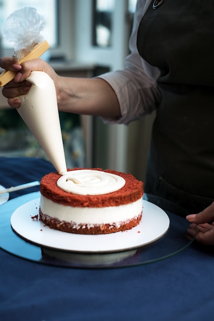 El pastelero exprime crema de color beige sobre pastel de terciopelo rojo. La mujer decora el pastel con crema dulce.