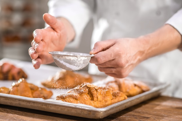 Foto pasteles dulces, decoración. manos de pastelero con un pequeño tamiz sobre productos horneados terminados, espolvoreando la superficie con azúcar en polvo