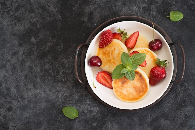 Pasteles de requesón con bayas y menta en un plato blanco sobre un fondo oscuro