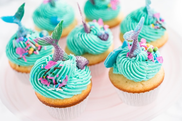 Pastelitos de sirena gourmet cubiertos con glaseado de crema de mantequilla azul y decorados con chispas y colas de sirena de chocolate.