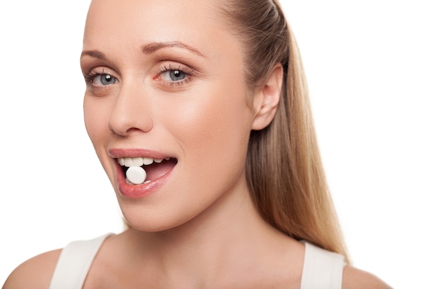 Pastilla en su boca. Retrato de mujer joven sosteniendo la píldora en la boca y sonriendo mientras aislado en blanco