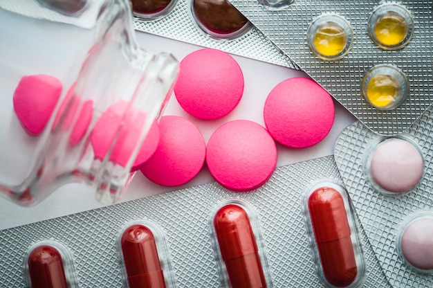 Las pastillas de color rosa se vierten de un frasco de vidrio y las ampollas con tabletas de colores se dispersan sobre un fondo blanco. La vista desde la cima.