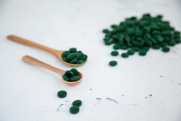 Pastillas de espirulina verde sobre un fondo de hormigón blanco. Varias pastillas en cucharas de madera