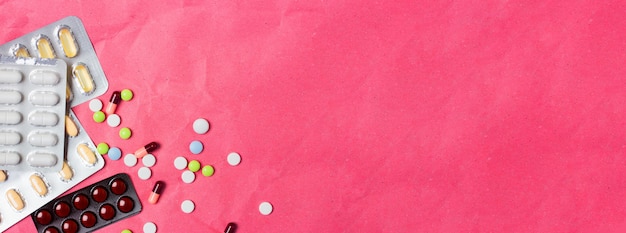 Pastillas multicolores y gránulos en una ampolla se encuentran dispersos sobre un fondo rosa. Concepto médico banner vista superior. Deslice el lugar para el texto.