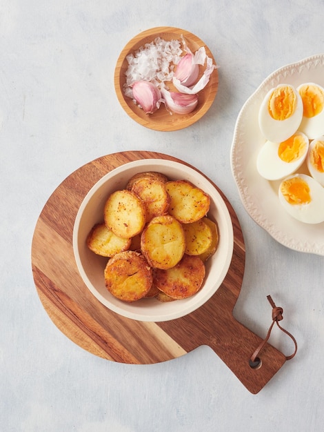 Patatas asadas crujientes de color marrón dorado cortadas junto a un plato con mitades de huevo duro Sal marina gruesa y dientes de ajo crudos en un pequeño plato de madera