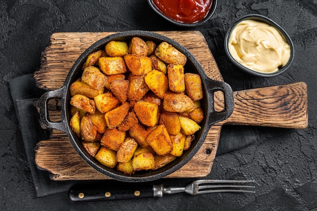 Patatas Bravas, würzige Kartoffeln, ein spanisches Gericht mit Bratkartoffeln und einer würzigen Knoblauchsauce. Schwarzer Hintergrund. Ansicht von oben.