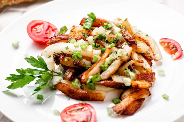 Patatas fritas en un plato blanco con cebollas verdes, perejil, tomates sobre un fondo de madera blanca.