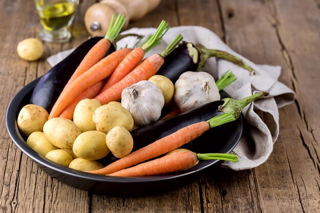 Patatas jóvenes crudas Zanahoria Ajo y berenjena en plato negro Variedad de verduras crudas