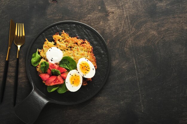 Patatas latkes con crema agria ensalada de espinacas tomates y huevos cocidos sobre fondo de mesa vieja de madera oscura Tortitas caseras de patata Deliciosa comida para el desayuno Vista superior