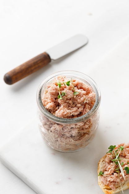 Paté de atún saludable o rillettes en un frasco de vidrio sobre tablero de mármol y fondo blanco