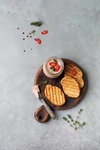 Paté casero de pollo de hígado con pimiento rojo, ramitas de tomillo y rebanadas de pan tostado sobre tabla vieja de madera sobre hormigón gris o piedra.