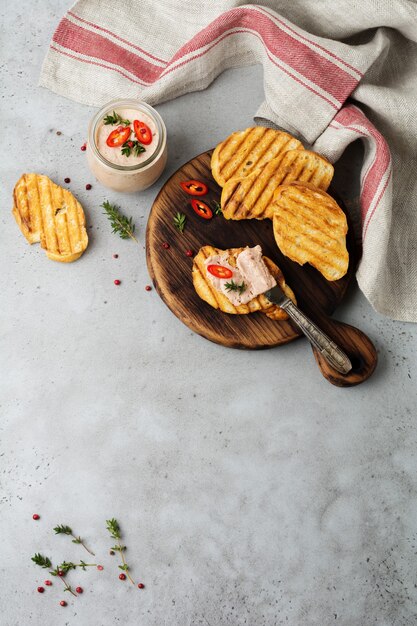 Paté casero de pollo de hígado con pimiento rojo, ramitas de tomillo y rebanadas de pan tostado sobre tabla vieja de madera sobre hormigón gris o piedra.