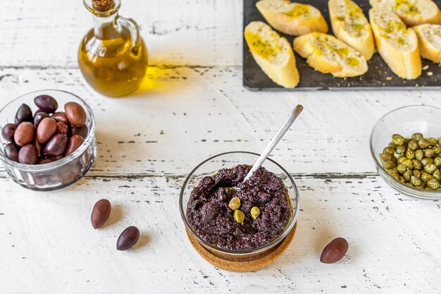 Patê de azeitona na tigela e ingredientes básicos azeite azeitonas e alcaparras em fundo branco de madeira