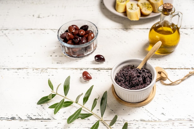 Paté de oliva en tazón de ramequin, aceite de oliva, aceitunas sobre fondo blanco de madera