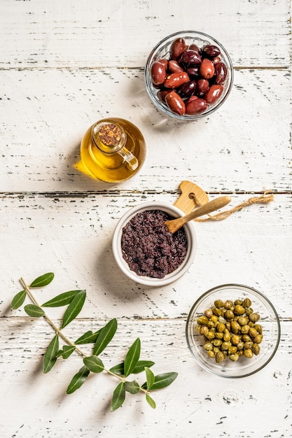 Paté de oliva en tazón de ramequin e ingredientes básicos sobre fondo blanco de madera con rama de olivo