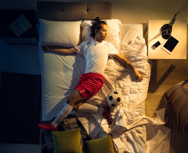 Pateando. Vista superior del fútbol profesional joven, jugador de fútbol durmiendo en su habitación en ropa deportiva con pelota. Ama su deporte, adicto al trabajo, juega partidos incluso si está descansando. Acción, movimiento, humor.