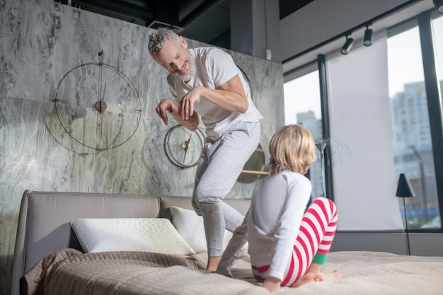 Paternidad. Papá barbudo canoso en pijama entretenido niño mostrando divertidas figuras en la cama en casa