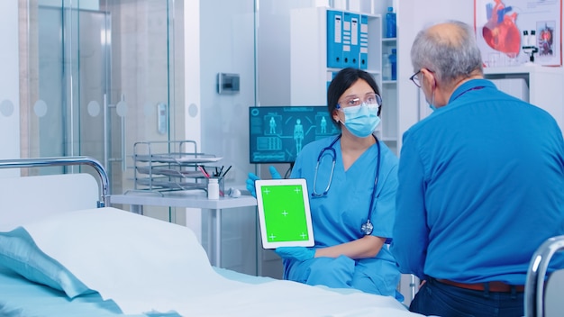 Patient, der ein digitales Tablet mit grünem Bildschirm in einem modernen privaten Krankenhaus oder einer Klinik betrachtet. Isolierter Mockup-Chroma-Ersatzbildschirm auf dem Gadget für Ihre App, Ihren Text, Ihr Video oder Ihre digitalen Assets. Easy Keying Medici