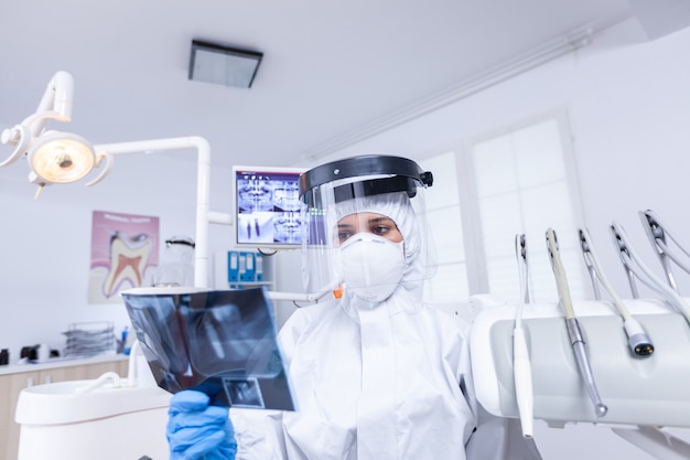 Patient pov in zahnarztpraxis, der zahnarzt mit röntgenaufnahmen in schutzausrüstung während covid-19 betrachtet. Zahnärztin, die einen Schutzanzug gegen Coroanvirus trägt und Röntgenaufnahmen zeigt