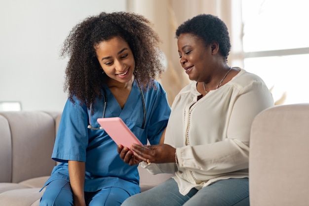 Patient und Krankenschwester. Dunkelhäutiger angenehmer Patient und Krankenschwester sitzen auf dem Sofa, während sie einen Film auf dem Tablet ansehen