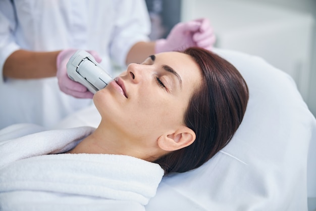 Patientin entspannt sich während der Ultraschall-Gesichtsbehandlung, die von einer Kosmetikerin in sterilen Handschuhen durchgeführt wird