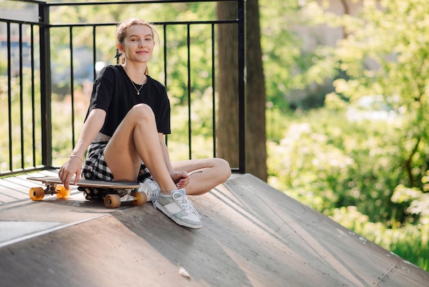 Una patinadora adolescente con una patineta está sentada en la rampa en el parque de patinadores