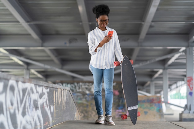 Patinadora africana com longboard segurar telefone e ler notificação de redes no espaço para skatistas