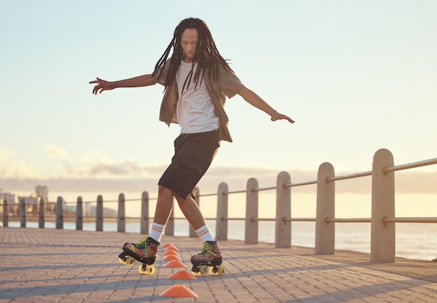 Patines en la playa y el deporte con un hombre cabalgando alrededor del cono de entrenamiento para divertirse y hacer ejercicio en el paseo marítimo Hipster atleta masculino patinando al aire libre para el ocio, la salud y la recreación