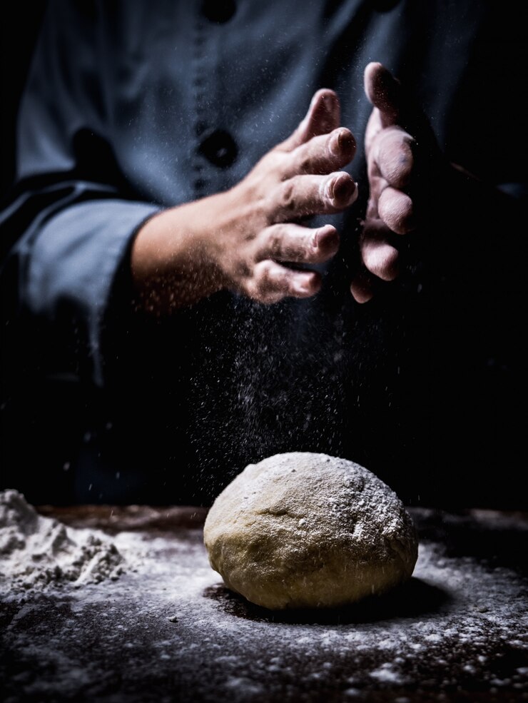 Patissierhand, die Weißmehl über rohem Teig auf Küchentisch besprüht.