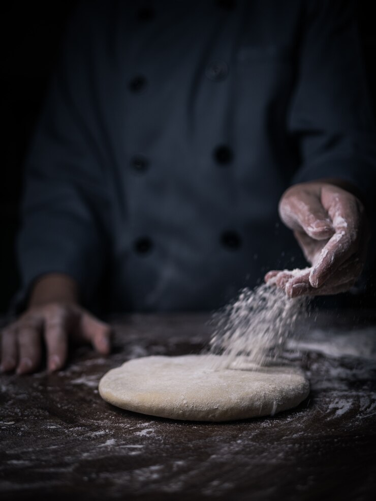 Patissierhand, die Weißmehl über rohem Teig auf Küchentisch besprüht.