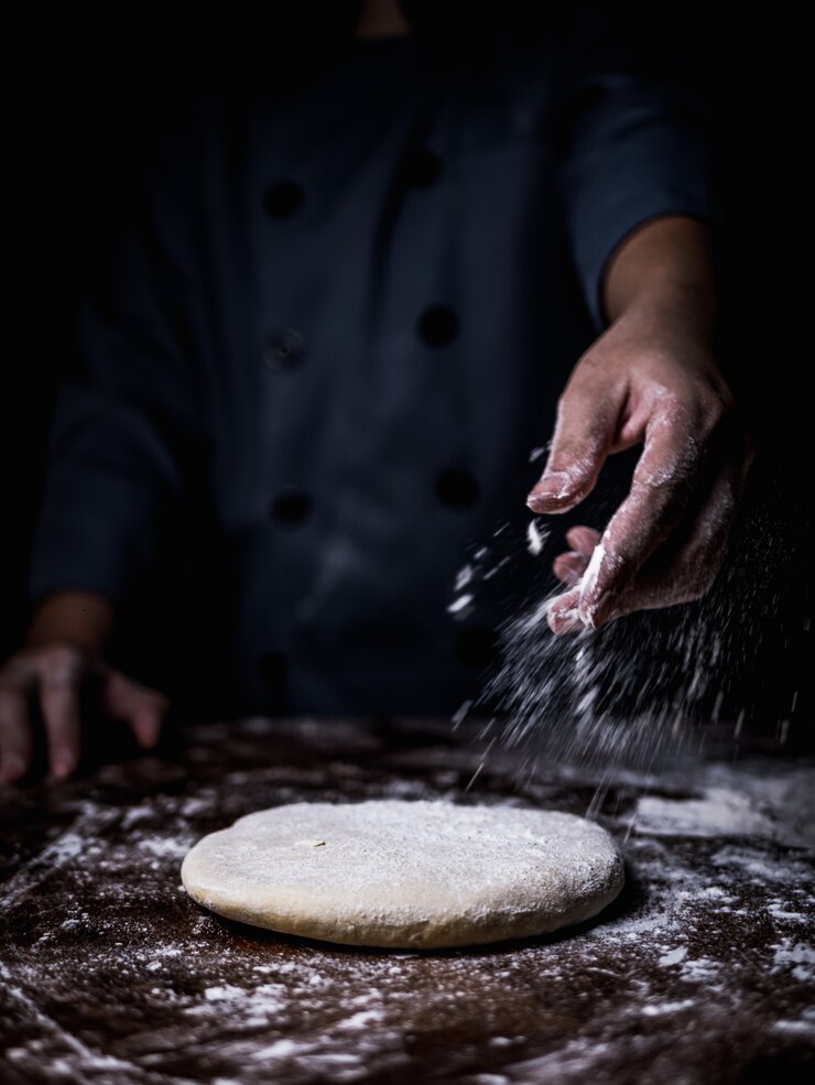 Patissierhand, die Weißmehl über rohem Teig auf Küchentisch besprüht.