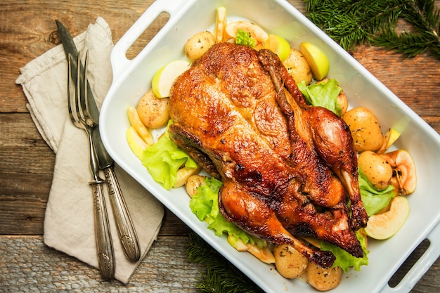 Pato de Navidad con manzanas y hierbas en sartén blanca sobre fondo de madera, vista superior