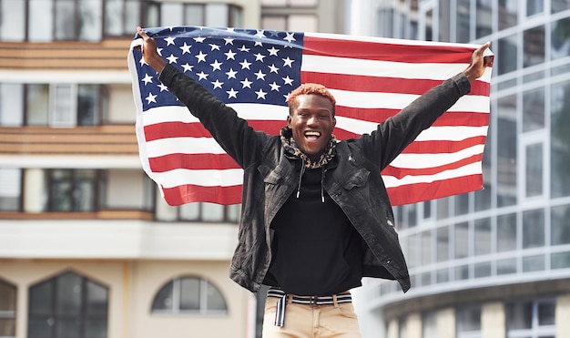 Patriot, der die USA-Flagge hält Konzeption von Stolz und Freiheit Junger afroamerikanischer Mann in schwarzer Jacke im Freien in der Stadt, der gegen ein modernes Geschäftsgebäude steht