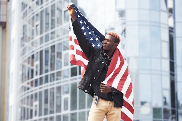 Patriota sosteniendo la bandera de Estados Unidos Concepción de orgullo y libertad Joven afroamericano con chaqueta negra al aire libre en la ciudad de pie contra el edificio de negocios moderno