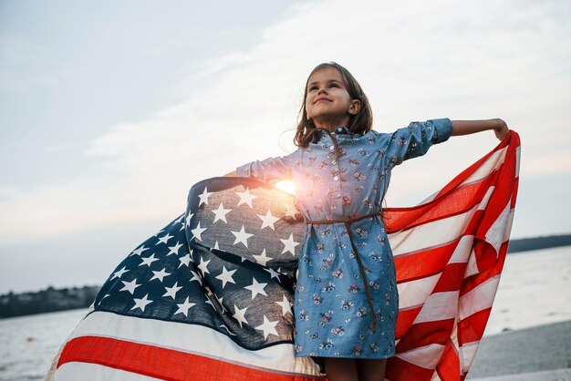 Patriotisches weibliches Kind mit amerikanischer Flagge in den Händen gegen bewölkten Himmel