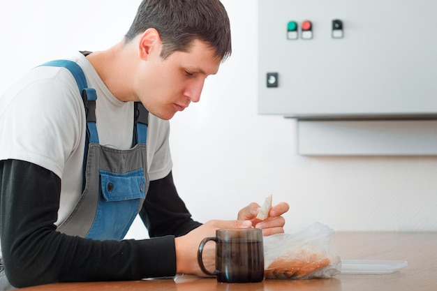 Pausa para o almoço Comendo no local de trabalho durante o trabalho Homem caucasiano em traje de trabalho senta-se à mesa na sala de produção e come do contêiner Trabalhador almoça na sala de descanso