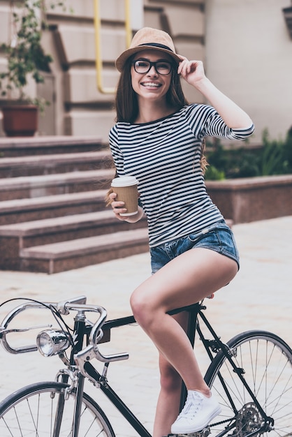 Pausa para o café antes do passeio. Mulher jovem e bonita segurando a xícara de café e se inclinando em sua bicicleta com um sorriso ao ar livre