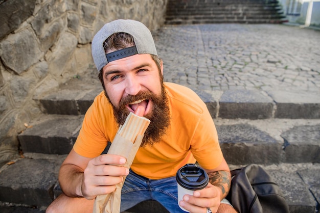 Pausa para salchicha. Chico caucásico con descanso para comer en el fondo urbano. Hombre barbudo con hot dog y café para la merienda. Hipster comiendo bocadillos en las escaleras durante el descanso.