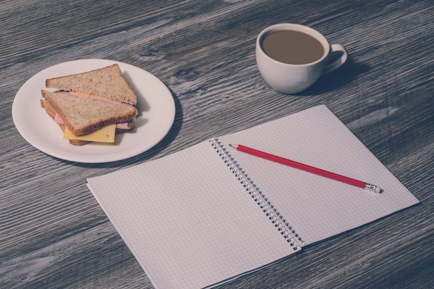 Pause bei der Arbeit. Offenes Schreibheft mit Bleistift, Schinken und Käsesandwich auf einem weißen Teller, Tasse heißen Tee auf Holzhintergrund. Vintage-Effekt