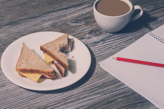 Pause bei der arbeit. sandwich mit gebissenem schinken und käse auf einem weißen teller, tasse heißen weißen kaffees, offenes notizbuch mit bleistift darauf auf holztisch. hintergrund, vintage-effekt