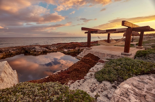 Pavillon mit zwei Hängematten darunter, Naturpool und Sonnenuntergang: ein Bild, das pure Entspannung anregt. Der Blick auf die private Küste von Puerto Aventuras in Mexiko.